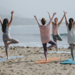 group of women performs standing yoga poses by the beach